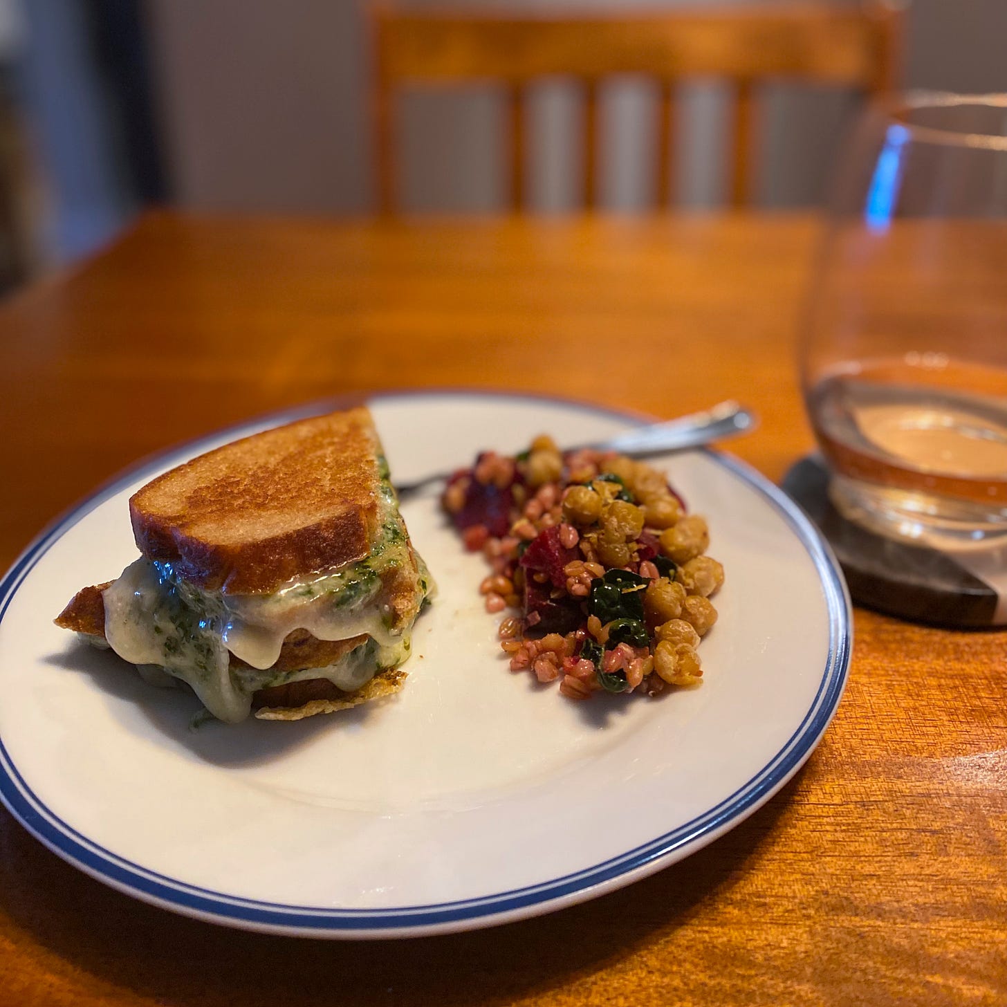 A plate with two halves of a grilled cheese with pesto. Some of the cheese has spilled out at the edge, parts of it browned and crisp. Next to the sandwich is a small pile of the farro salad described above, and there is a glass of rosé wine on the coaster next to the plate.