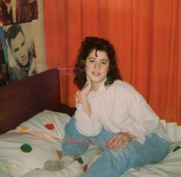 A teen me, brunette with a perm, wearing a baggy white shirt and blue jeans, is sitting on a bed infront of closed orange curtains. The duvet cover is white with colourful spots, and I am posing for the camera. There are posters of bands on my wall.