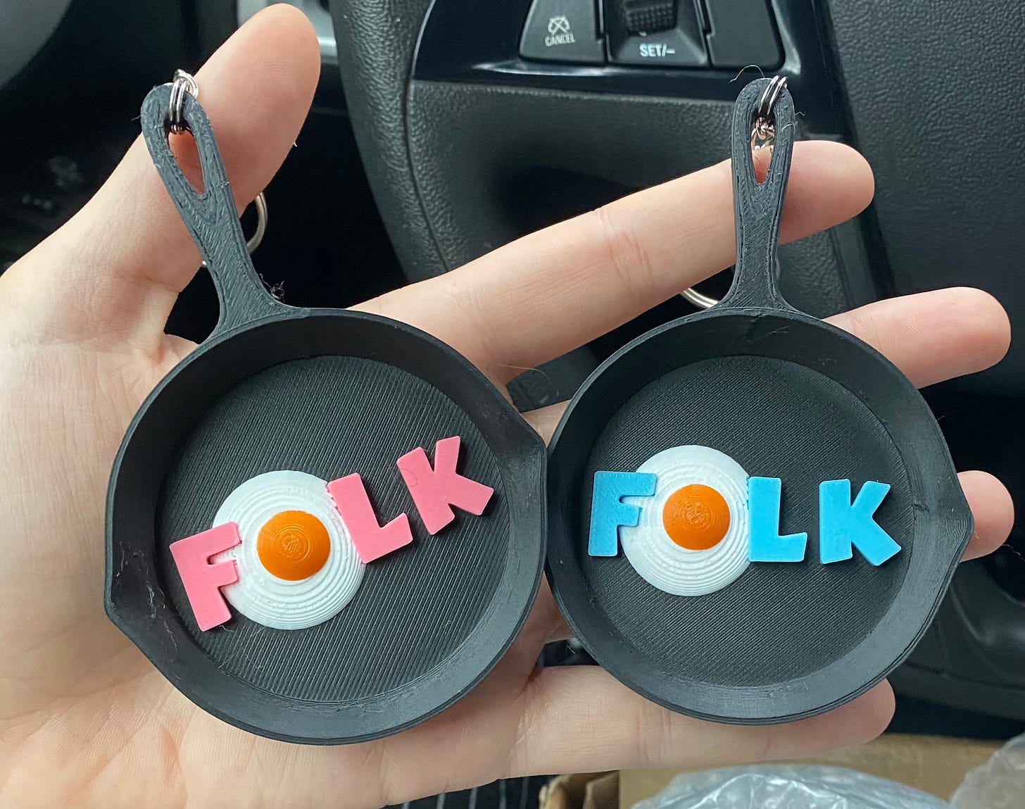 Shady's hand holding two mini 3D printed cast iron keychains with "FOLK" written inside them. The "O" is an egg.