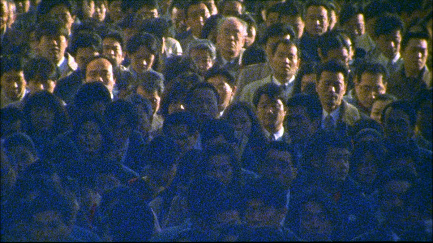 A crowd of people. Only their faces are visible, and the upper half is lit in yellow daylight. The bottom is in shadow, with a blue tinge over their faces.