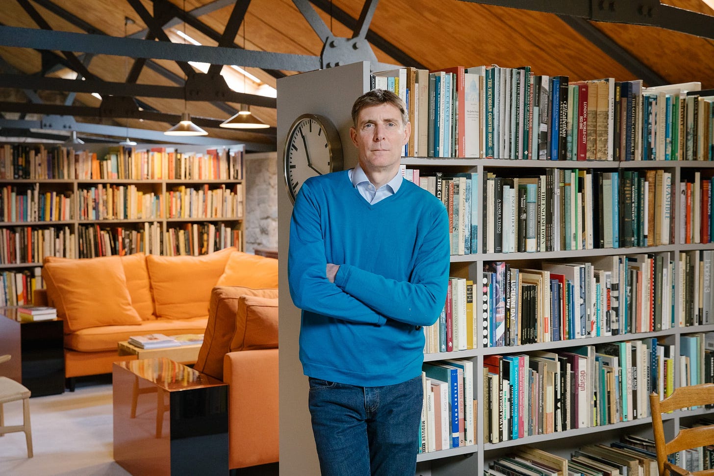 A portrait of Corin Mellor wearing a v-neck jumper and shirt. He is leaning on the corner of a bookcase with books also behind him.