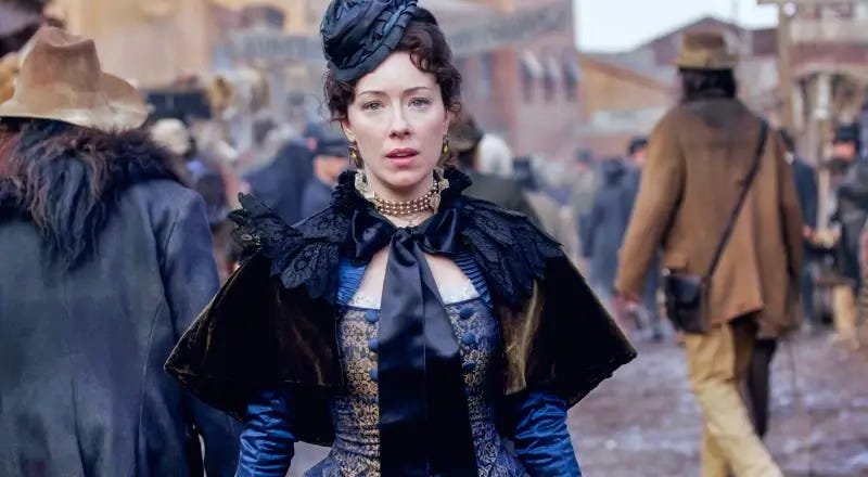 This image shows Alma Garret (played by Molly Parker) wearing a beautiful blue outfit as she stands in Deadwood's main thoroughfare and stares in shock at something or someone off-camera.