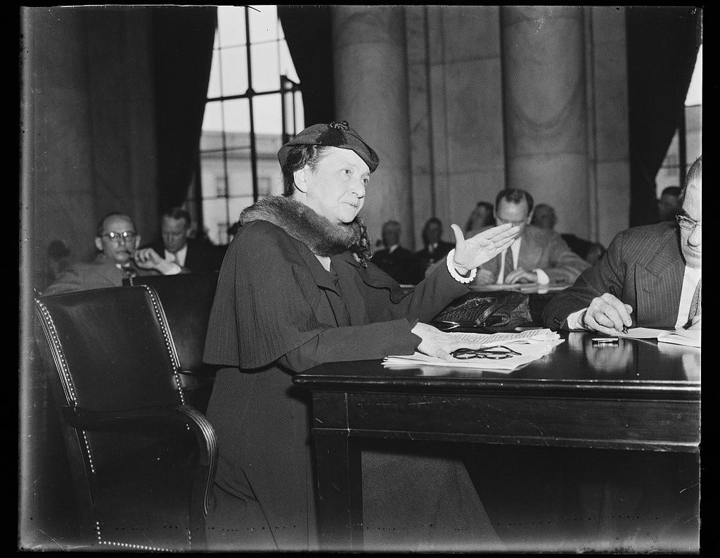 a black and white photo from 1936 shows Frances Perkins in a dark coat, hat, and fur stole seated at a table with papers in front of her. Men sit in an audience behind her