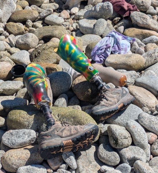 a pair of colorful tie-dye prosthetic legs with hiking boots at one end, plus other disassembled leg parts, laying on the rocks in the sunshine near a t-shirt and a towel