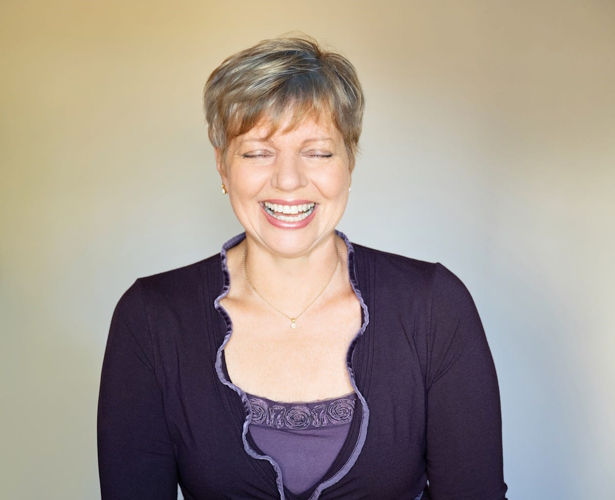 Woman laughing, eyes shut, wearing a purple top against a plain background.