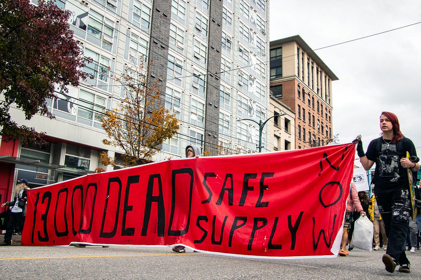 a large banner reads "13,000 dead. safe supply now" the background is red and the text is black. it is about three road lanes wide, and it's being carried by a group of people marching among a large crowd, which is largely unseen behind them