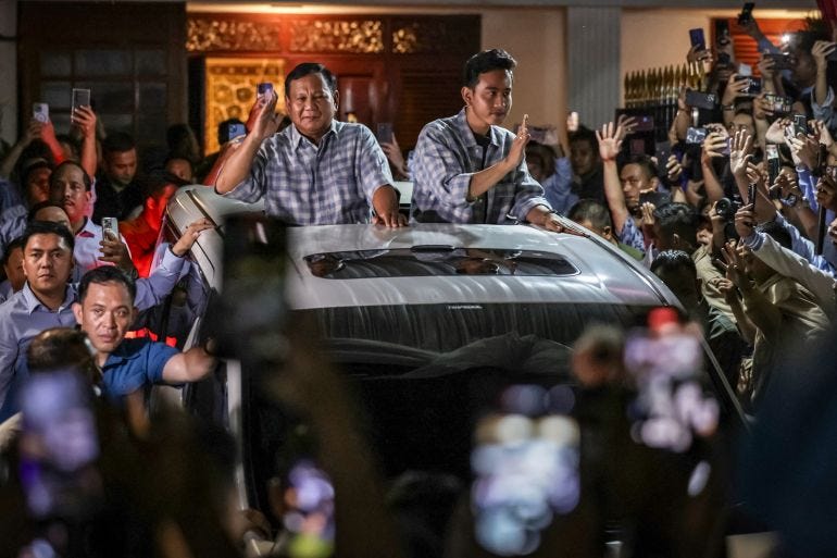 Prabowo and Gibran in an open top vehicle greeting supporters after quick count votes showed they'd won the 2024 election. There are crowds around the vehicle, which is flanked by security. Prabowo is saluting. Gibran is waving