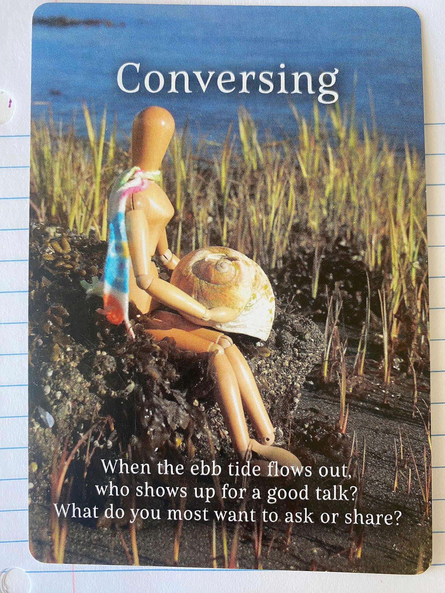 Supposing card of the Emotikin art manikin (inner artist) at the beach sitting next to a seashell. The card is titled Conversing and the caption says “When the ebb tide flows out, who shows up for a good talk? What do you most want to ask or share?” 