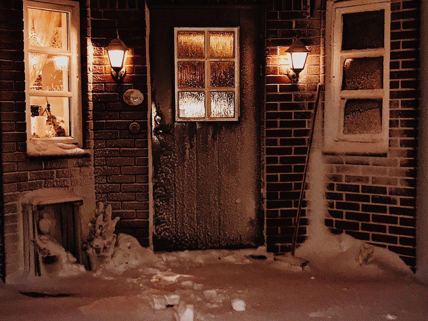 Brick building with windows and wall lights on either side of the front door. The ground and outside of the building are covered with snow. 