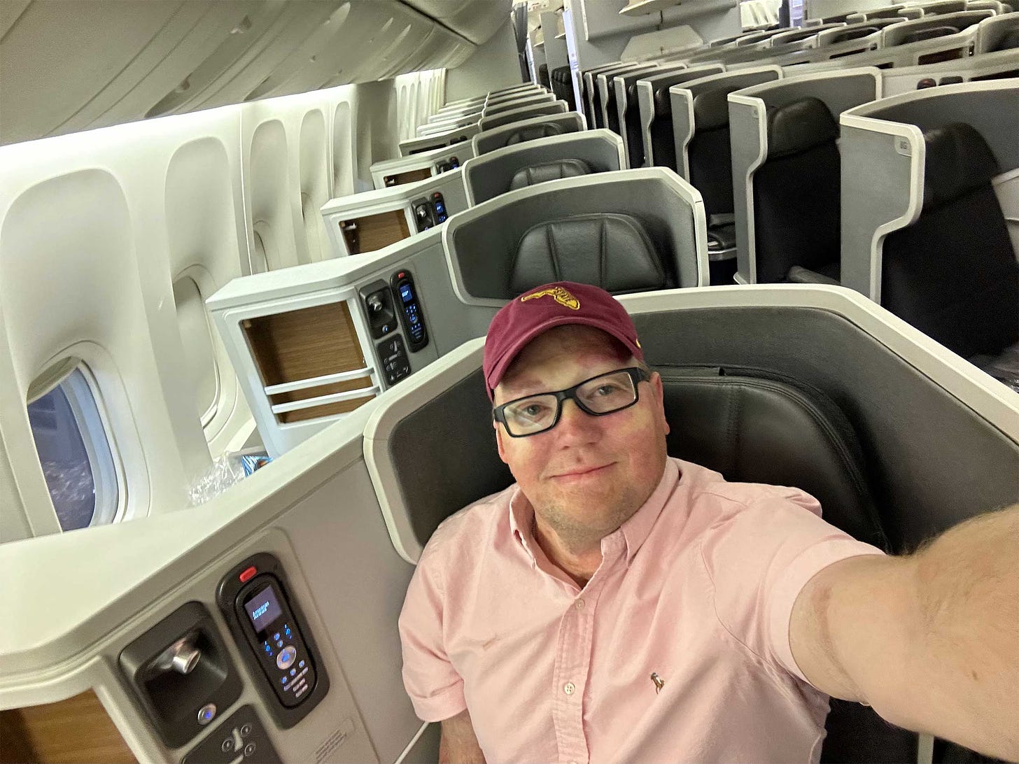 Selfie of John wearing a pink shirt and garnet baseball cap seated in business class on a Boeing 777 aircraft.