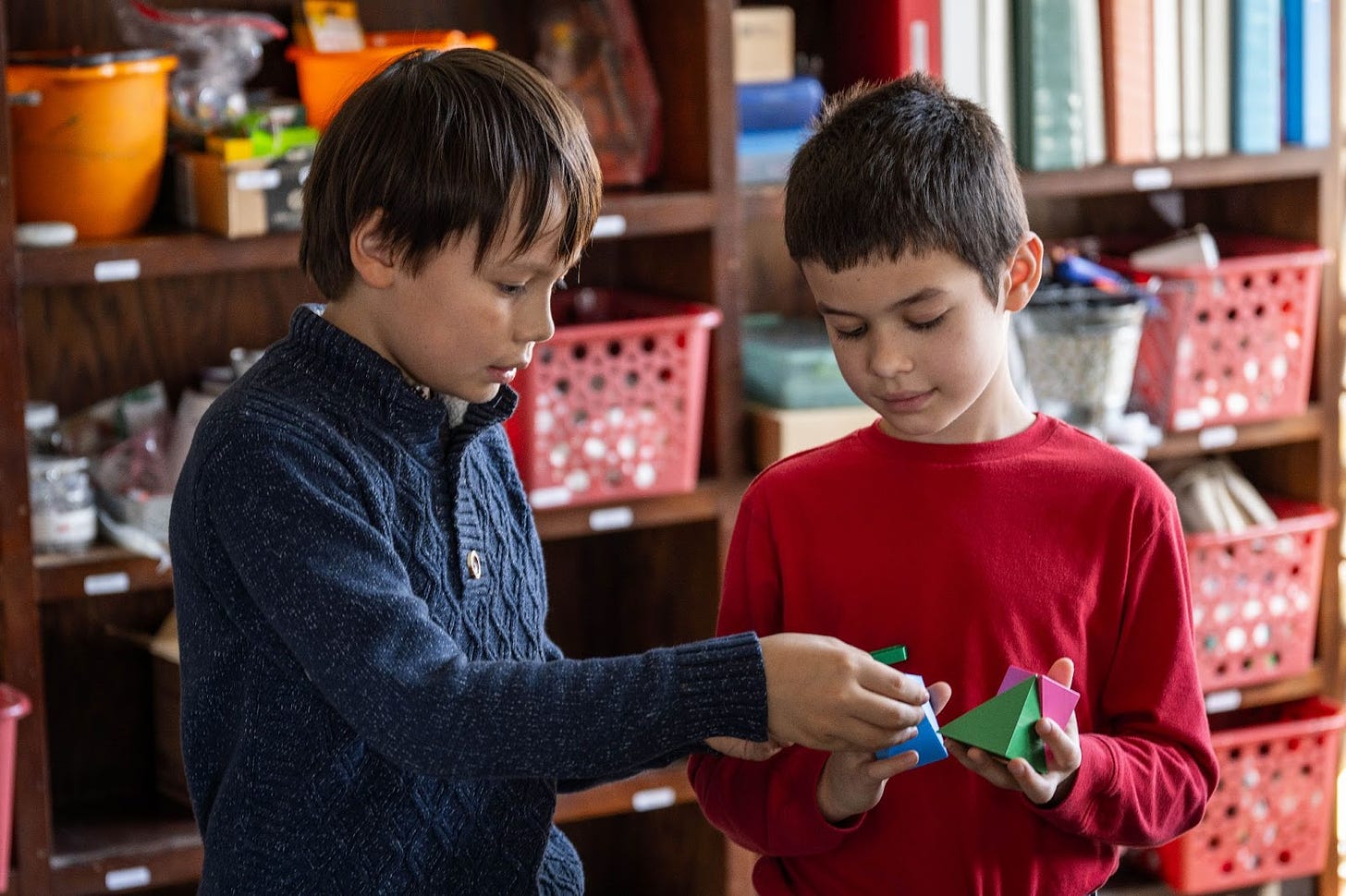 Two young boys seem to be in a school setting with baskets of supplies along the wall behind them. One boy is holding a partially-assembled 3D printed puzzle that makes a cube, and the other boy is helping put the last piece in place.