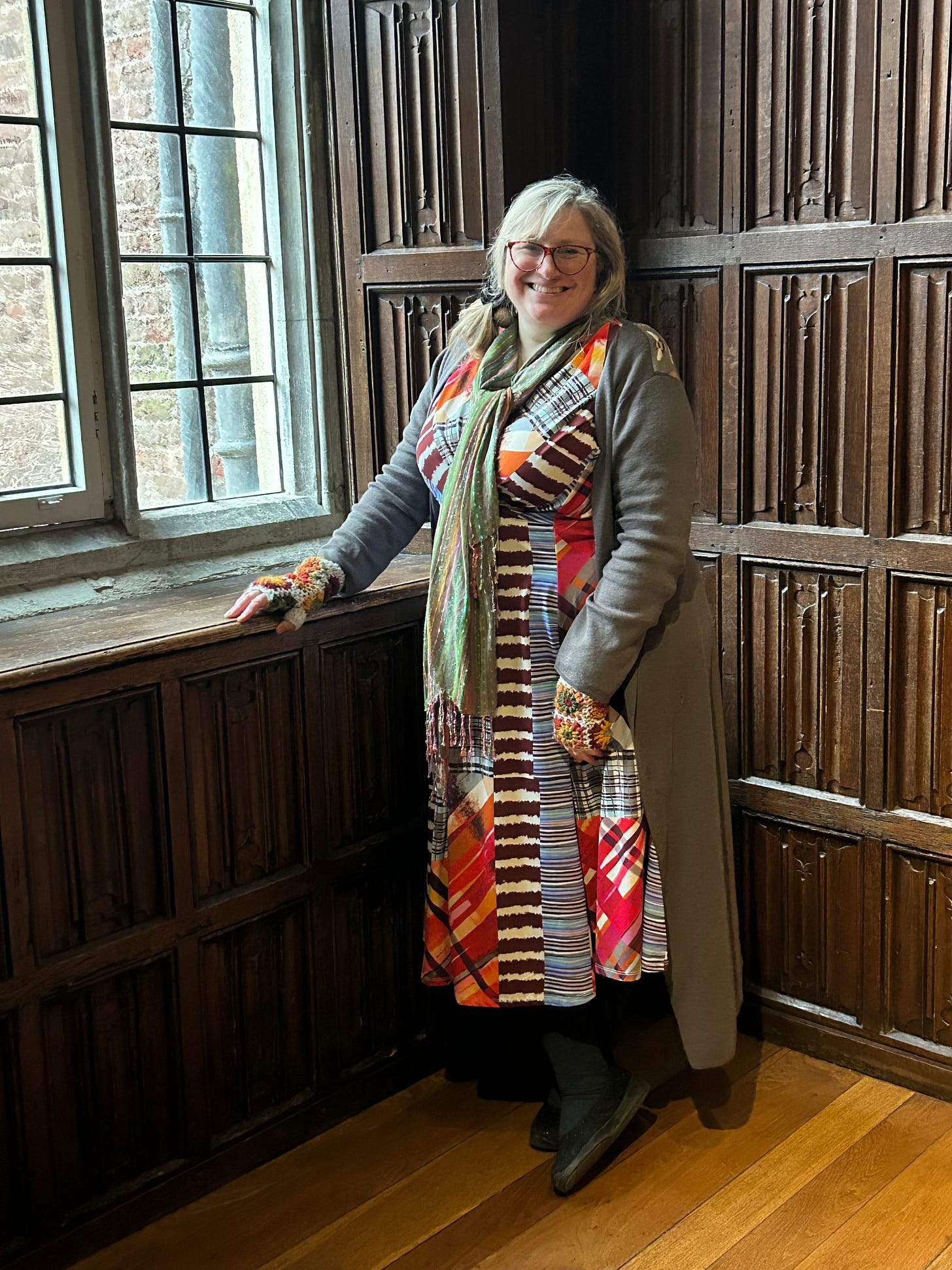A white woman wearing a brightly coloured dress stands in a wood panelled room next to a leaded window.
