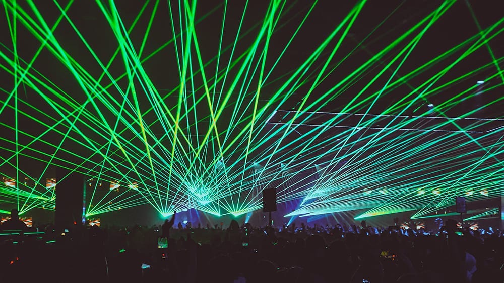 A large crowd of people dancing at a rave as green lasers shoot overhead