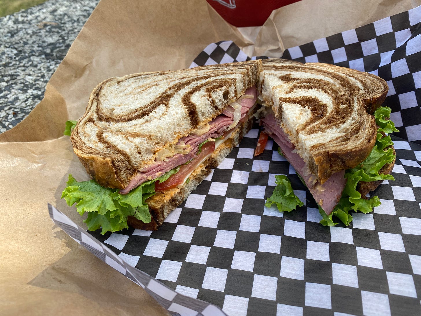 The reuben from Nature's Pickins, on marble rye bread, cut in half and sitting on checkered deli paper on a stone outdoor picnic table.