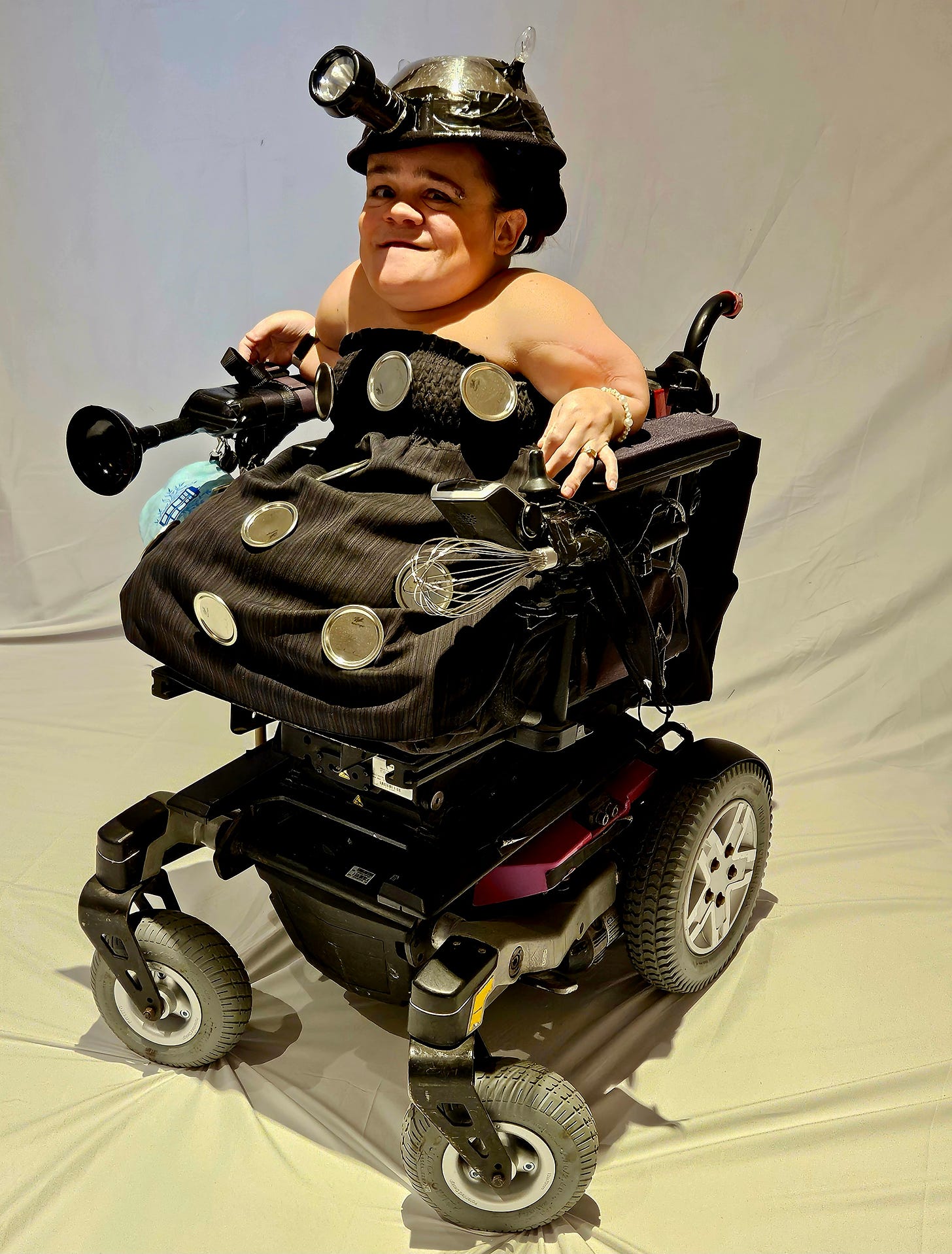 Costume consists of a mixing bowl on her head, with a flashlight attached to the front like an eyestalk. There is a black plunger on her left wheelchair armrest, a whisk attached to her right wheelchair armrest, and 12 silver jar lids attached to her black dress to represent the circular shapes on the Dalek's armor.