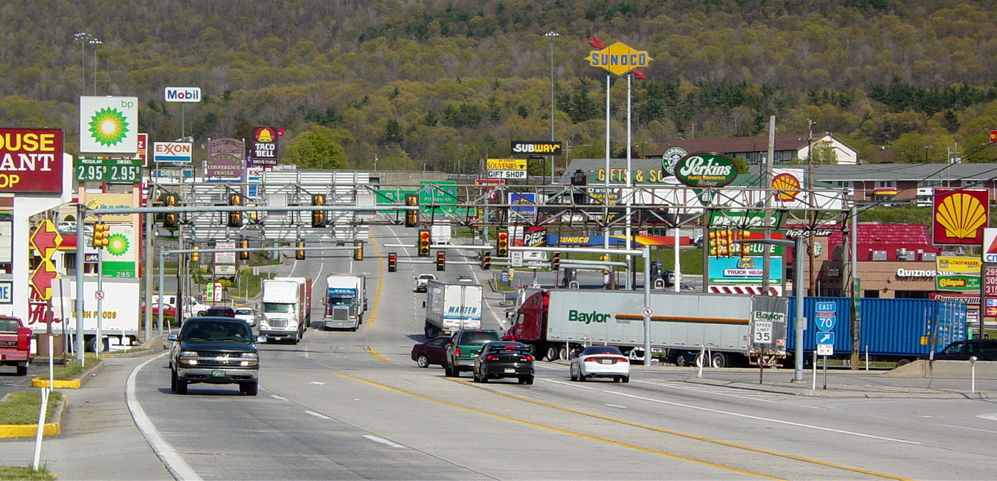 Image of a typical US highway interchange: a large five lane road with minimal pedestrian facilities with heavy trucks and lined with gas stations and fast food restaurants.
