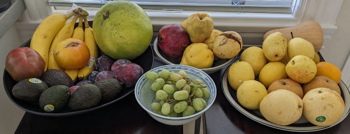 A display of several bowls overfilled with fruit, including bananas, tomatoes, avocados, grapes, pomello, pomegranate, quince, Asian pears, lemons, and a butternut squash