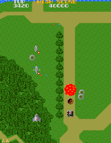 A screenshot of an early portion of Xevious, featuring an explosion on the ground, as well as various enemies. There's a road, grass, and trees all in sight, a huge departure for STGs that had, to this point, been all about mostly static black backgrounds with some stars in them.