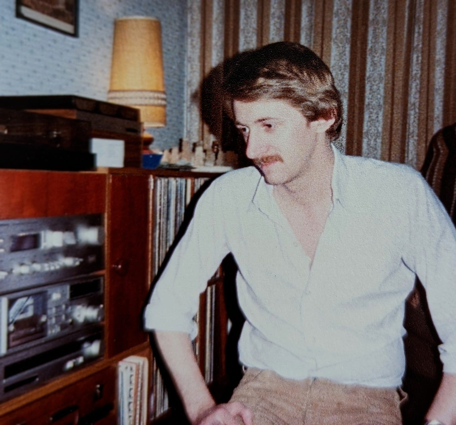 A young man with blonde hair and a moustache sits on the floor next to his beloved Cyrus hi-fi, LPs stacked on shelves. He is gazing to the side.