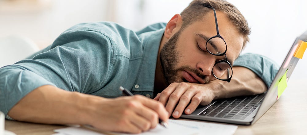 Stock photo of a man asleep, with his arm and head on his computer, his hand still in writing position, and glasses falling off his head. He appears to be falling asleep at his work desk.
