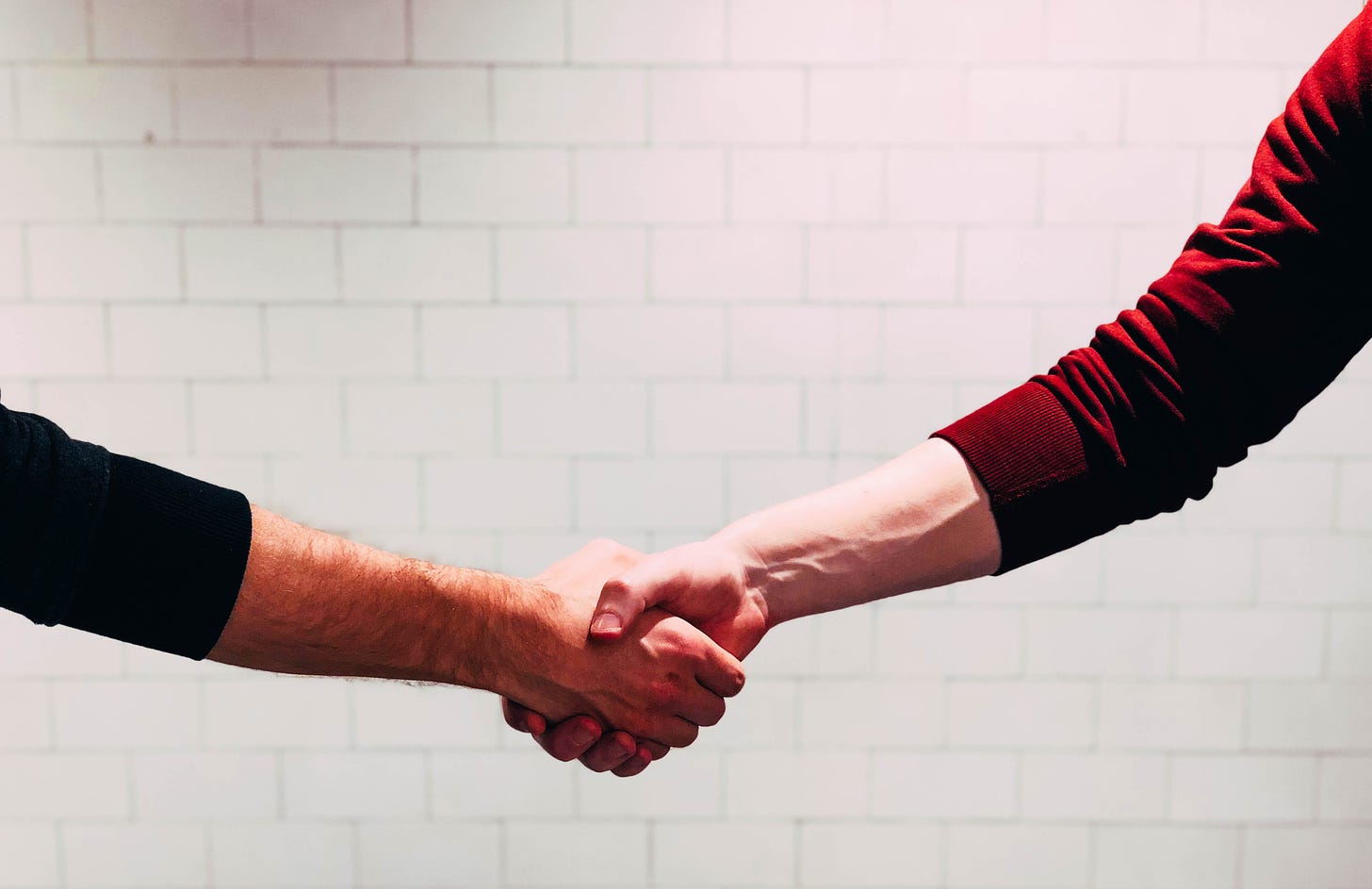 The arms of two caucasian people shaking hands against a white brick background. The arm on the left is wearing a black long-sleeved shirt, and the arm on the right is wearing a red long-sleeved shirt.