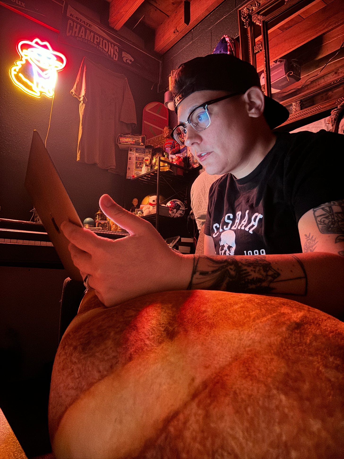 a white, masculine presenting person working on a computer atop a giant baguette pillow beneath many signs including a cat in a cowbow hat neon sign