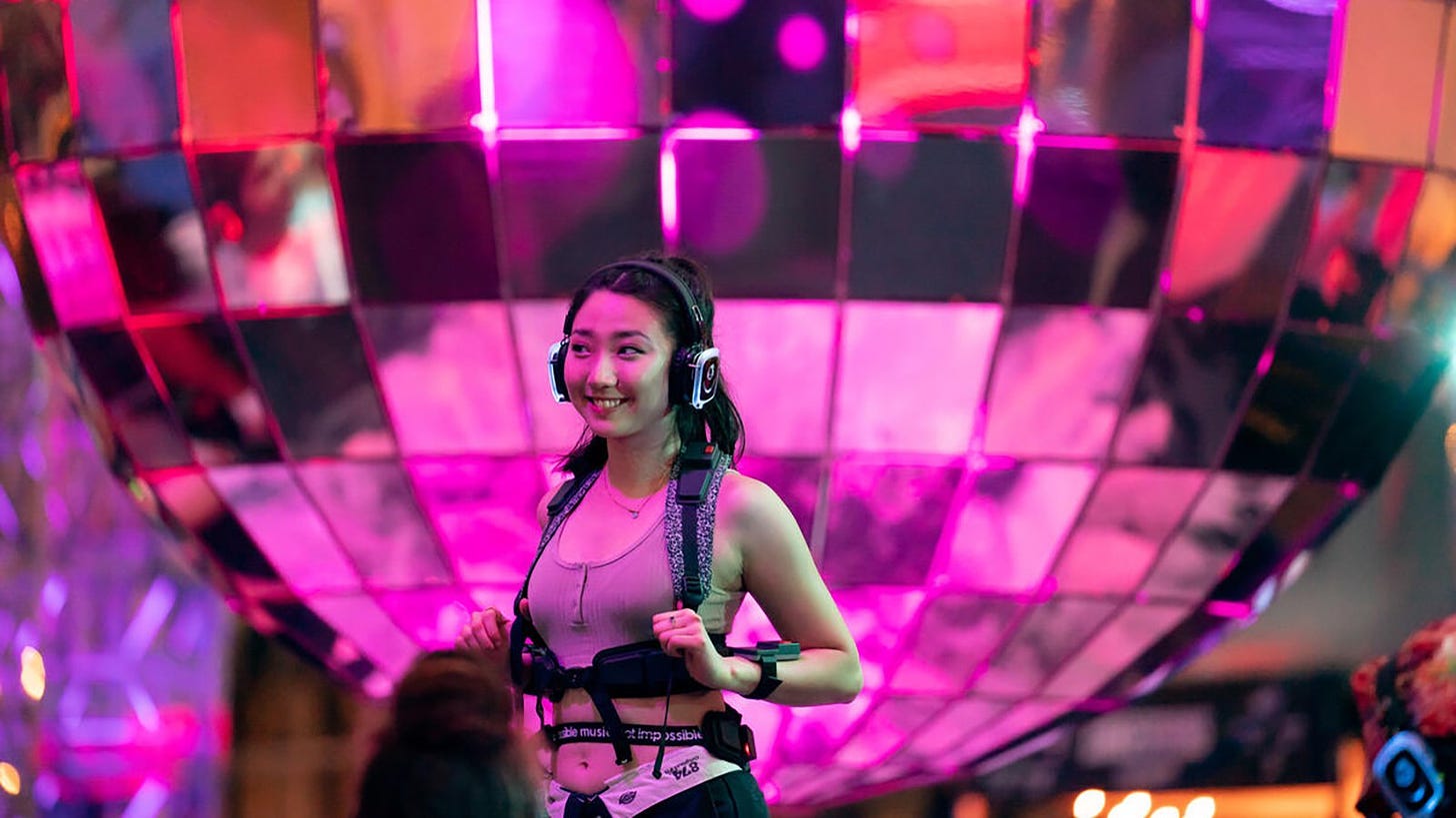 A party-goer is wearing headphones and a vest that pumps bass into the body, standing in front of a 10 foot tall disco ball at Lincoln Center.
