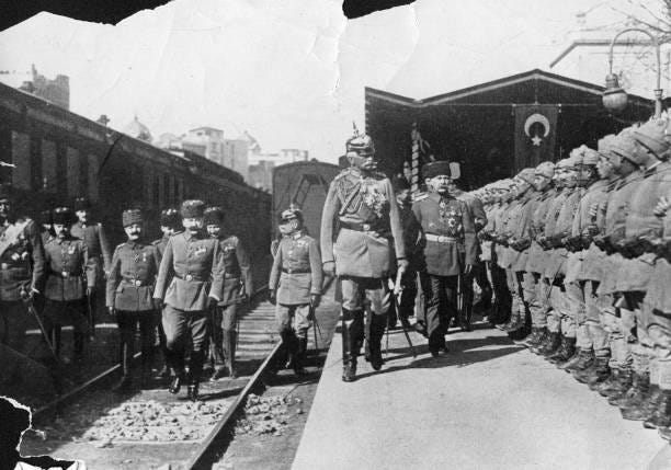 a man wearing a pickelhaube and a WWI era military uniform inspects a row of soliders at a railway station