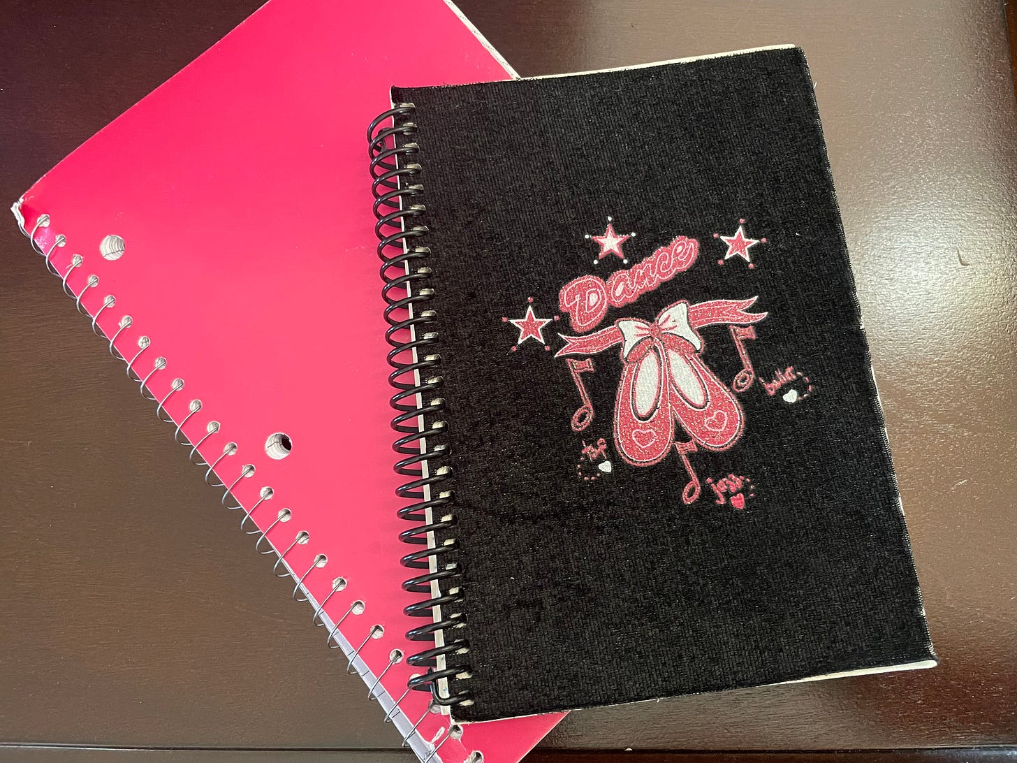 Two journals, one stacked on top of the other, atop a wooden table. The bottom one is slightly larger and pink. The top one is black, and says “Dance,” with pink illustrations of ballet shoes, hearts, and musical notes.