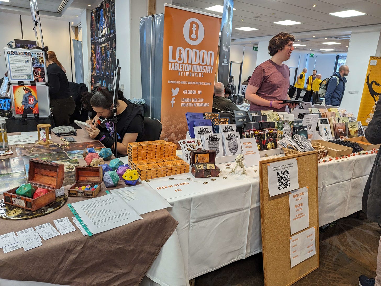 Michael Duxbury and Mikhail Malkin at the London Tabletop Industry Networking stall at Dragonmeet 2023. Visible on the table are various printed zine games on racks, character cards and dice-shaped candles.