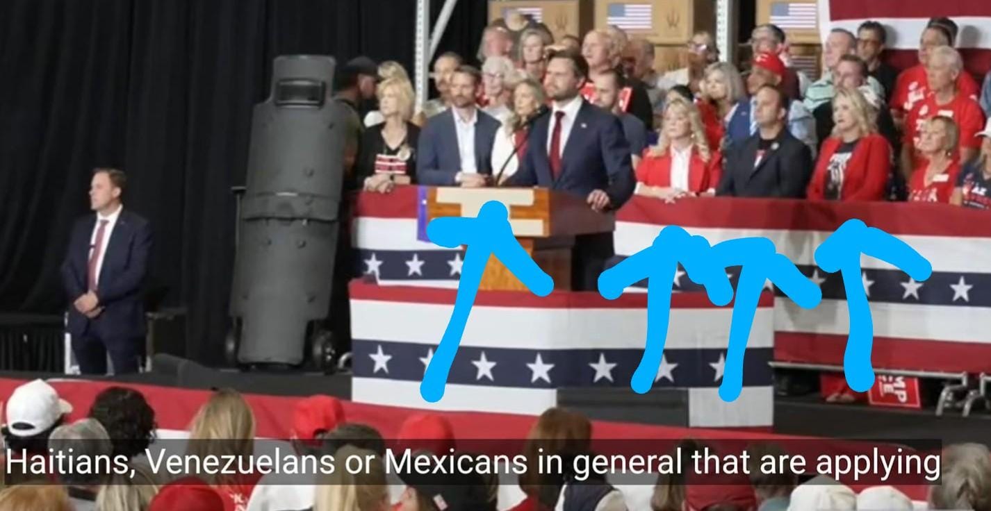 JD Vance stands at a podium at a campaign rally, surrounded by the Stars and Stripes. A row of very caucasian people sits on stage behind him. Blue arrows point up at them. Closed captions run across the bottom of the page, saying "Haitians Venezualans or Mexicans in general that are applying"