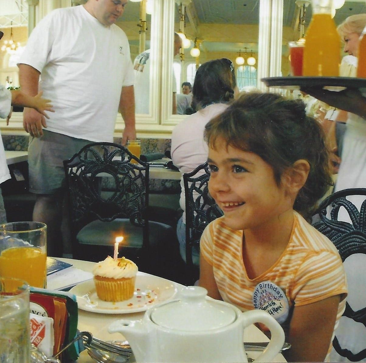 7-year-old girl sits by a cupcake with a birthday candle in it and smiles while others sing happy birthday to her