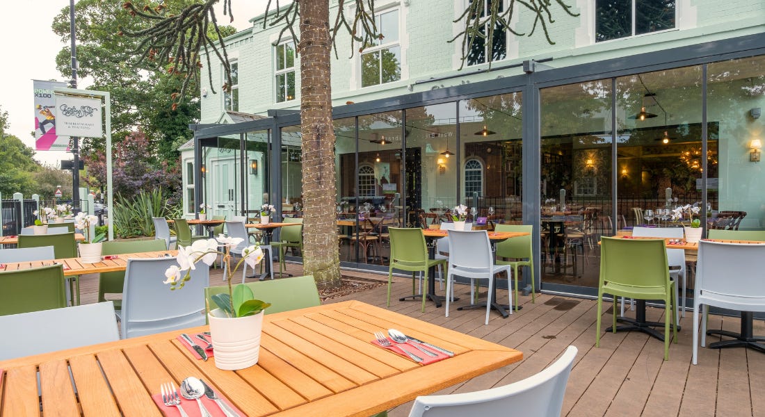 A green and brown outdoor dining area with wooden tables and green and grey chairs, and a mint green building to the right of it