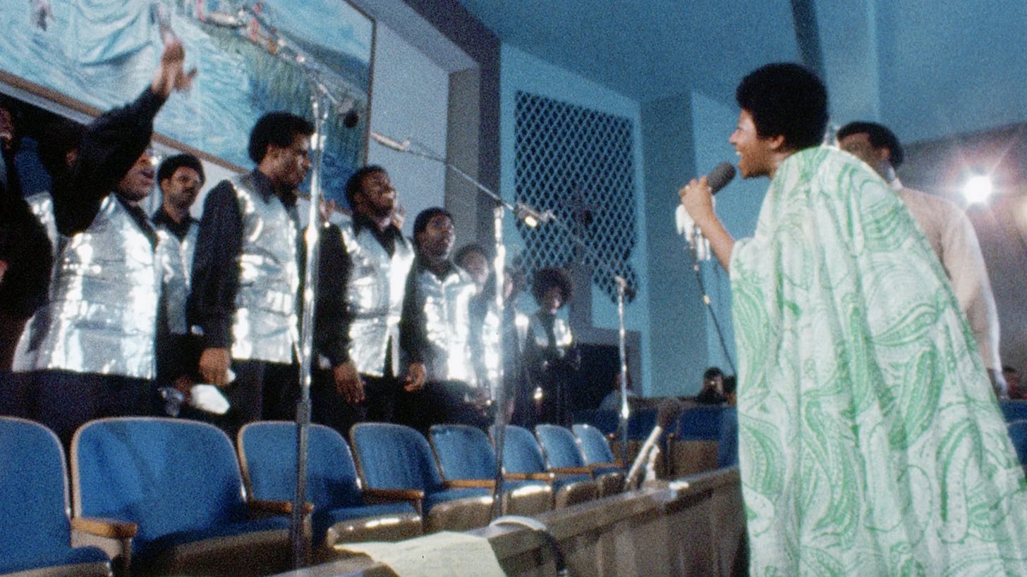 Aretha Franklin in a green dress singing to a choir wearing silver vests.