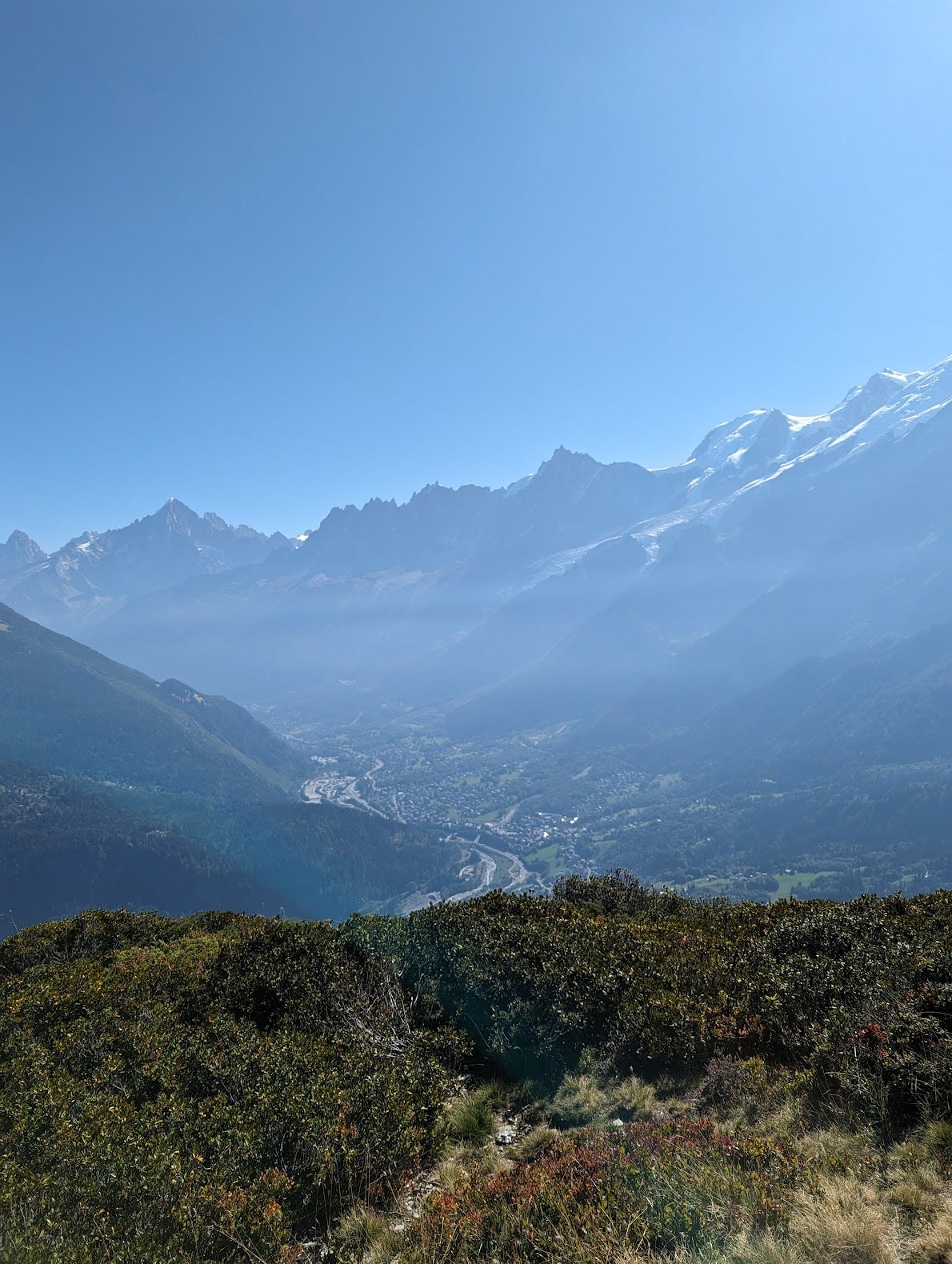 A photo taken from the mountains looking over Chamonix. The town appears small and far away, and mountains rise up along it on both sides.