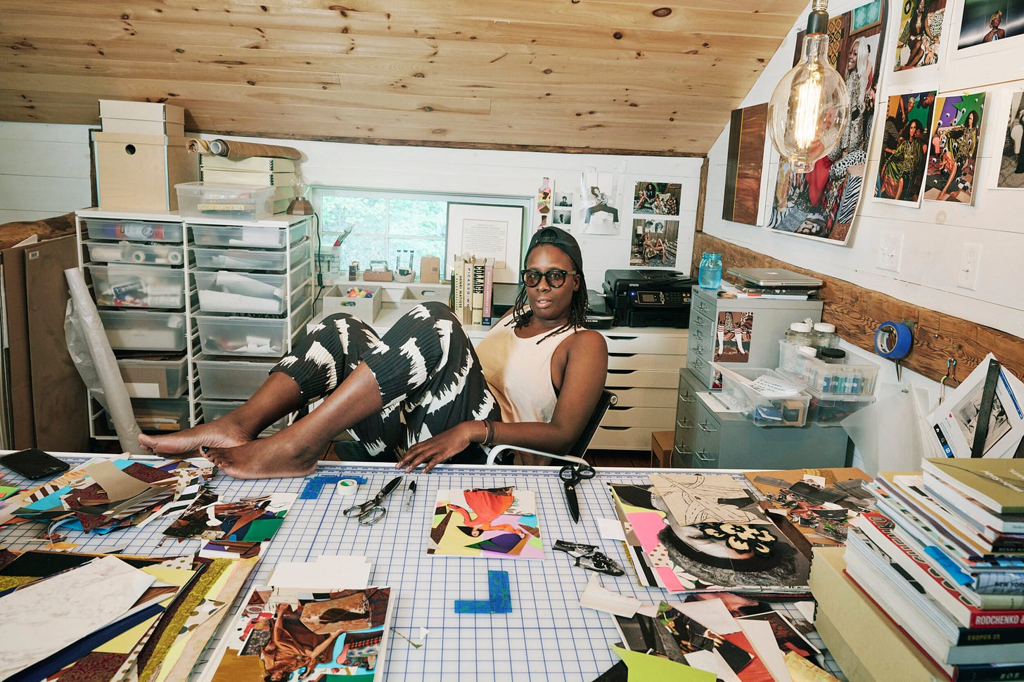 Mickalene is sitting on a chair in her collage studio. She has her feet up on the desk, and she is surrounded by scraps of paper, scissors, and paper clippings. The background contains more of her supplies, and the white wall is covered in her art. Mickalene is sitting on a chair in her collage studio. She has her feet up on the desk, and she is surrounded by scraps of paper, scissors, and paper clippings. The background contains more of her supplies, and the white wall is covered in her art.