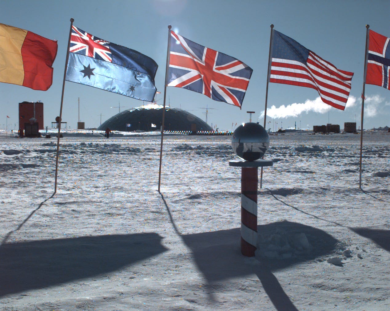 A photo taken in Antartica, with a blue partly cloudy sky over bright icy ground. In the background is the shiny dome of Amundsen-Scott Station. In the foreground is a short pole, red-and-white striped, with a shiny sphere on top, representing the south pole. Several flags fly behind the pole, all in a line. The ones in frame include Belgium, Australia, the UK, the US, and Norway.