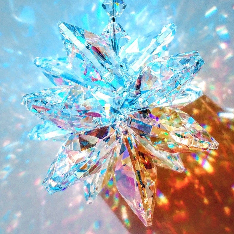 May include: A crystal starburst ornament with iridescent facets. The starburst is made of clear glass with rainbow reflections. The starburst is hanging from a silver string.