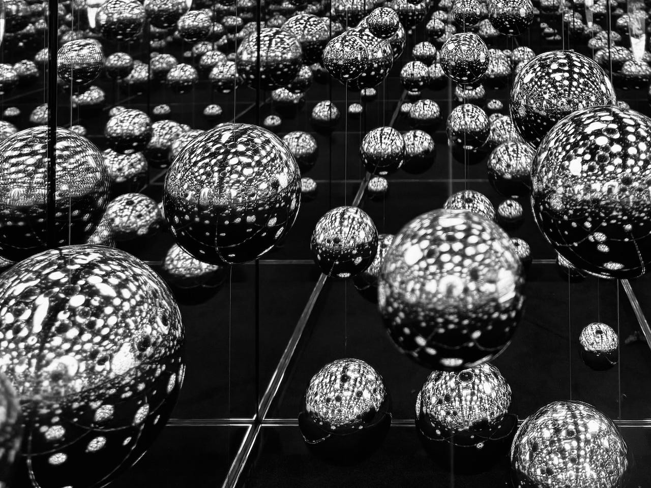 GPT-4o (edited): A black-and-white photograph of a mirrored space within an “infinity room” art installation, filled with mirrored spheres of various sizes. The reflective balls appear to be of different sizes, with the mirrored walls creating endless reflections of them, giving an illusion of infinite depth. The reflections of white light from circular openings add contrast and emphasize the intricate patterns on the mirrored surfaces. GPT-4o (edited): A black-and-white photograph of a mirrored space within an “infinity room” art installation, filled with mirrored spheres of various sizes. The reflective balls appear to be of different sizes, with the mirrored walls creating endless reflections of them, giving an illusion of infinite depth. The reflections of white light from circular openings add contrast and emphasize the intricate patterns on the mirrored surfaces.