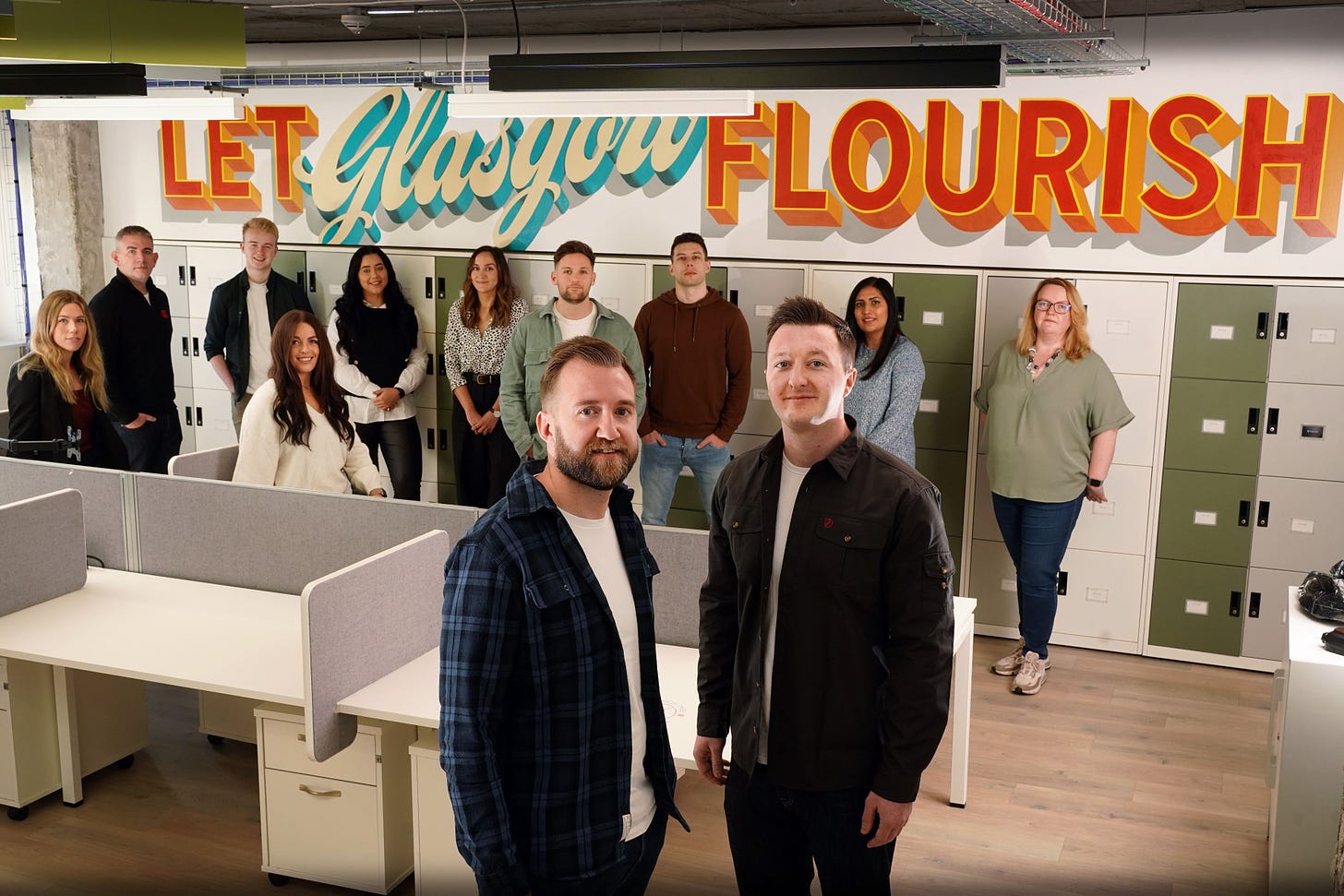 The twelve members of the Gigged.AI team, standing in an office, with founders Rich and Craig in the foreground. A mural on the wall behind the team reads 'Let Glasgow Flourish' 