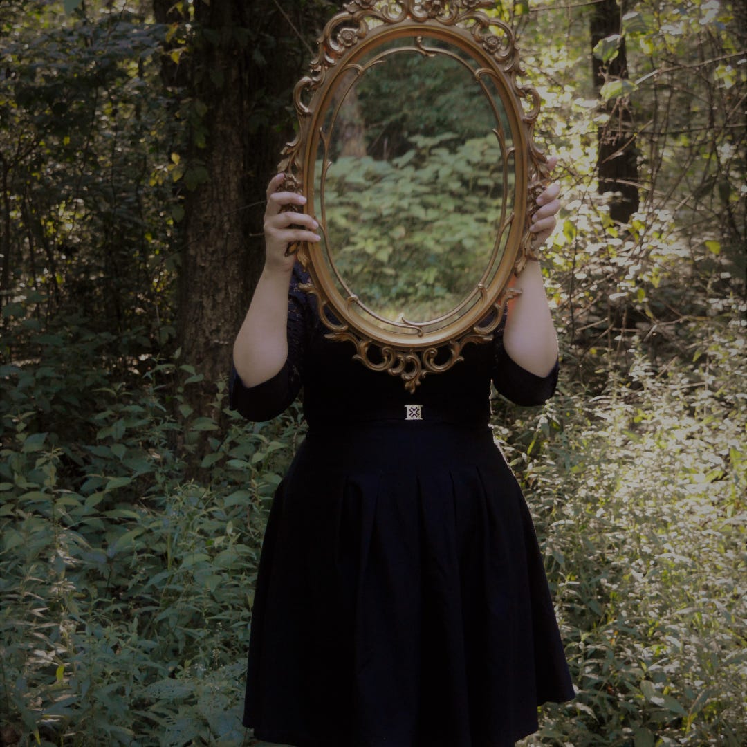 A figure dressed in black stands in a wood. They are holding a gold-framed mirror in front of their face which reflects the forest greenery all around them.