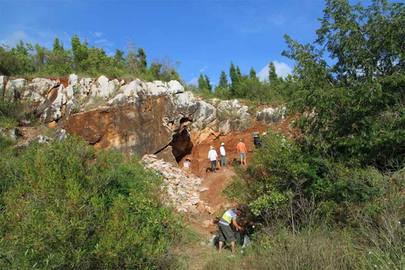 A rock wall with a cave and red sediments with archaeologists examining the site