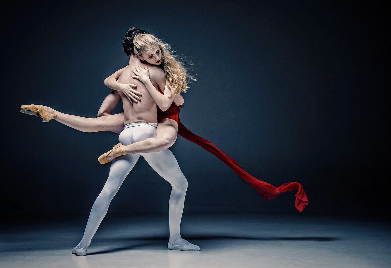A man and a woman are dancing ballet. He wears white tights and faces away from the camera in a slight lunge position. The woman is in his arms, clinging to him with her arms and one leg as her other leg points out behind him. The picture seems to have been taken just after she has landed the pose, as a stream of red silk is flowing out into the air behind her.