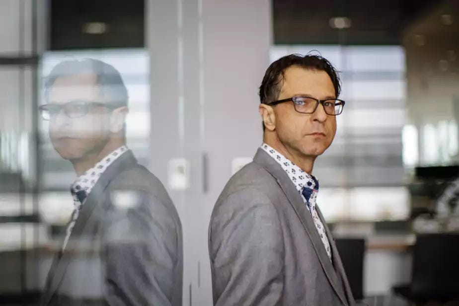 Carl Latulippe standing in front of some glass, looking to the side and looking right at the camera.