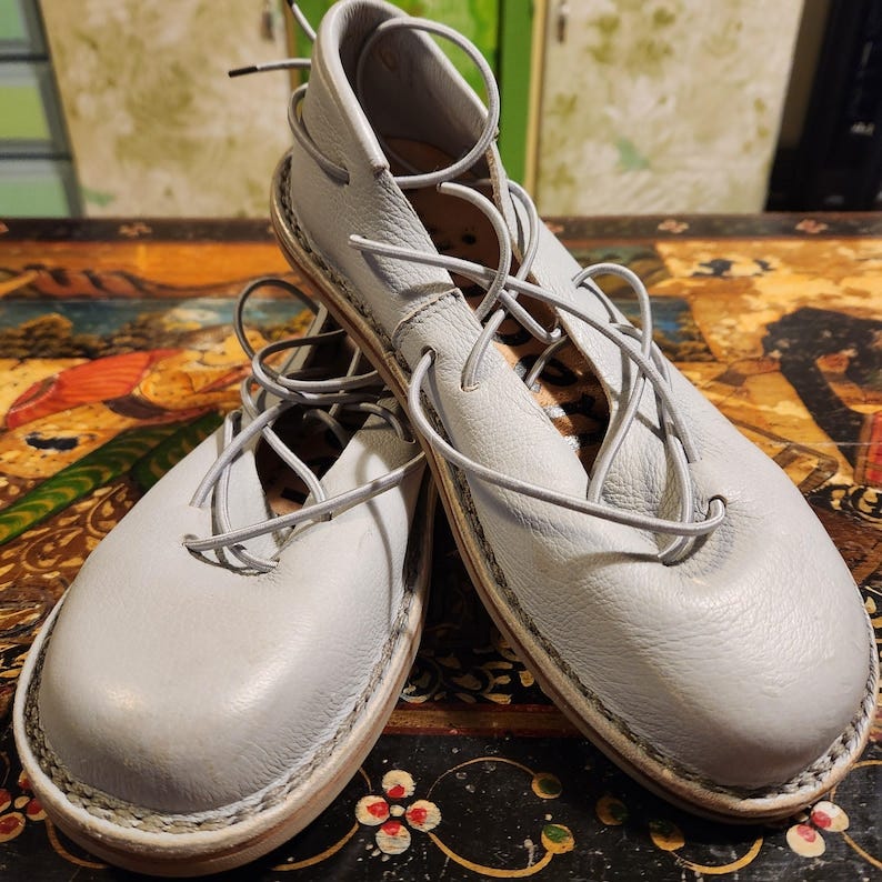 May include: Pair of light blue leather lace-up shoes. The shoes have a round toe and are adorned with laces that wrap around the ankles. The shoes are handcrafted with visible stitching and a light-colored sole. The shoes are displayed on a dark, ornate surface.