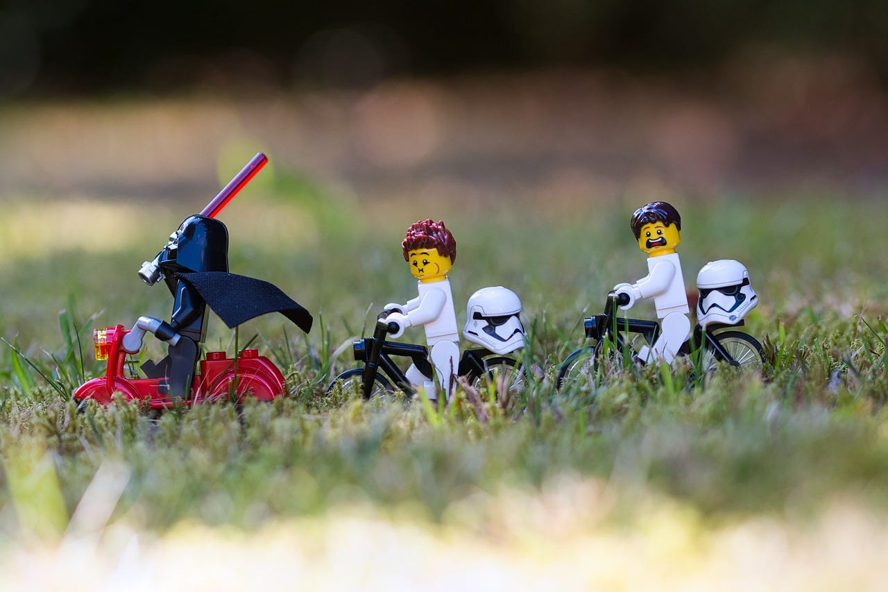 Three lego star wars figures riding bikes through the grass. Darth V. is first, followed by two storm troopers. 