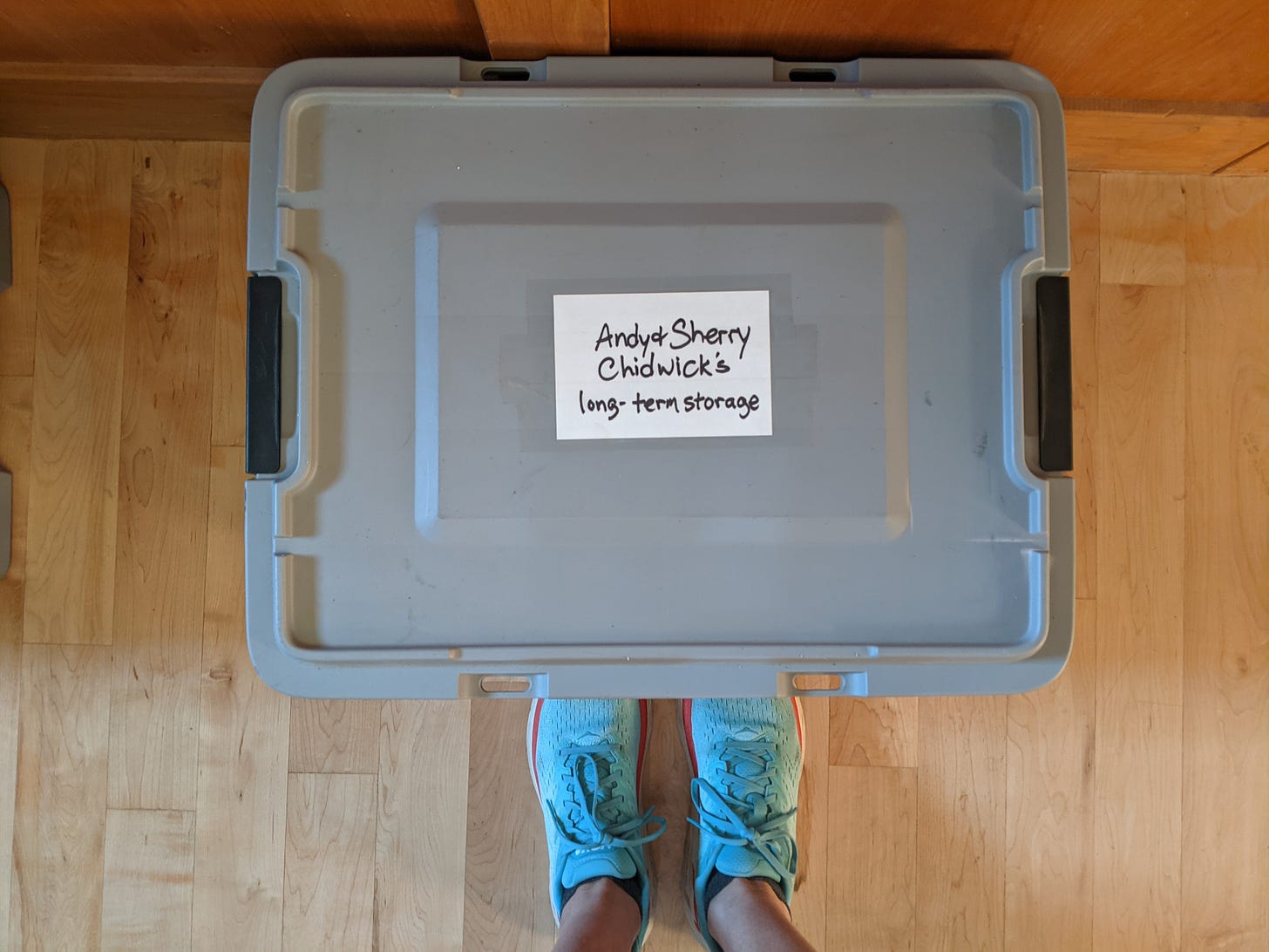 plastic tub labeled Andy & Sherry Chidwick's long-term storage, sitting on a hardwood floor in front of a pair of feet in blue running shoes