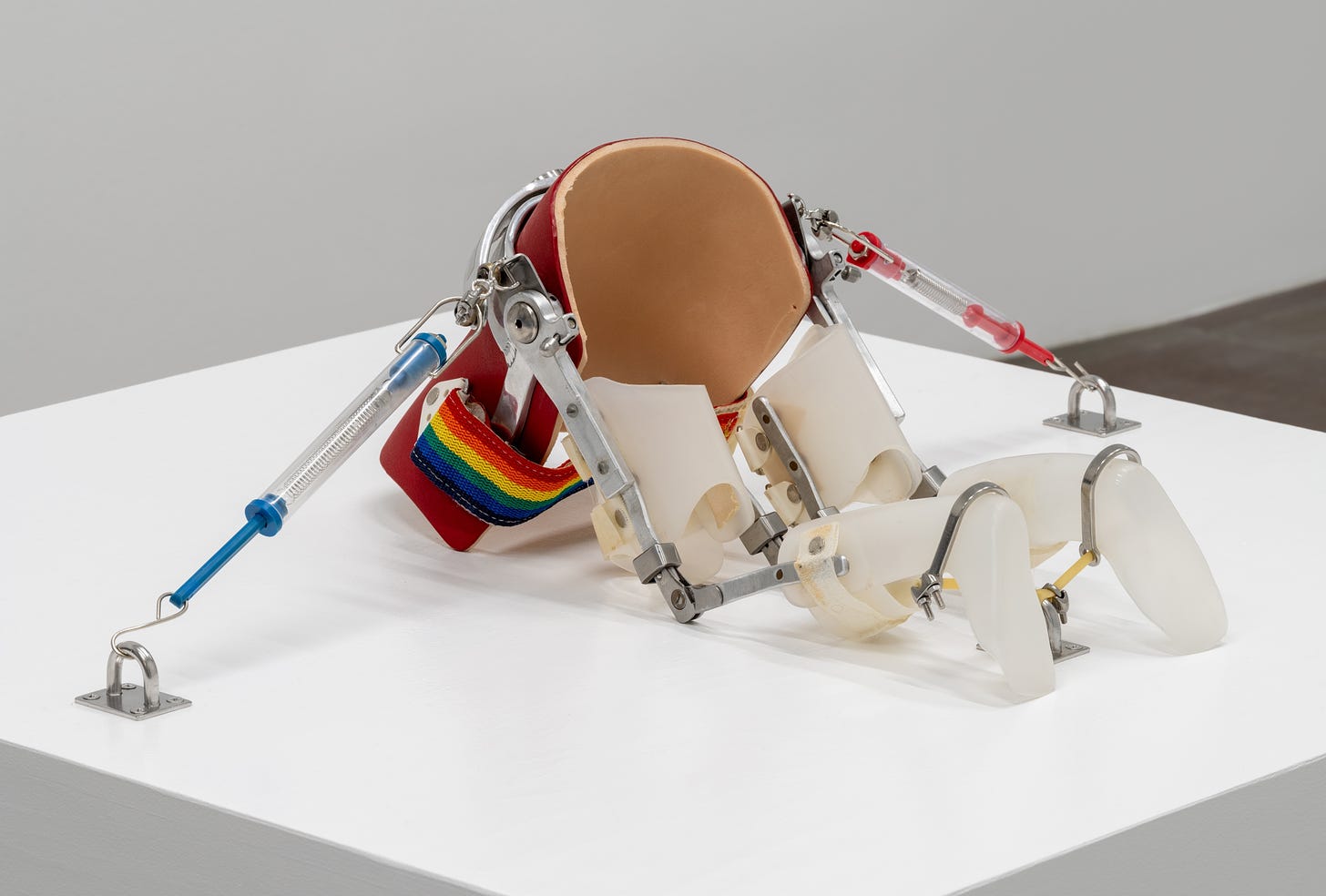 A photograph of a sculpture installed on a white plinth in a gallery. A body is formed by negative space. Miniature leg braces form 2 legs, attached to a back brace pointed downward so it looks like the body is kneeling and the upper half is missing or disappears into the plinth. A blue and a red syringe-like gauge form the arms, hooked on metal hooks mounted to the base. There is a stretchy rainbow strap that would cross the belly.
