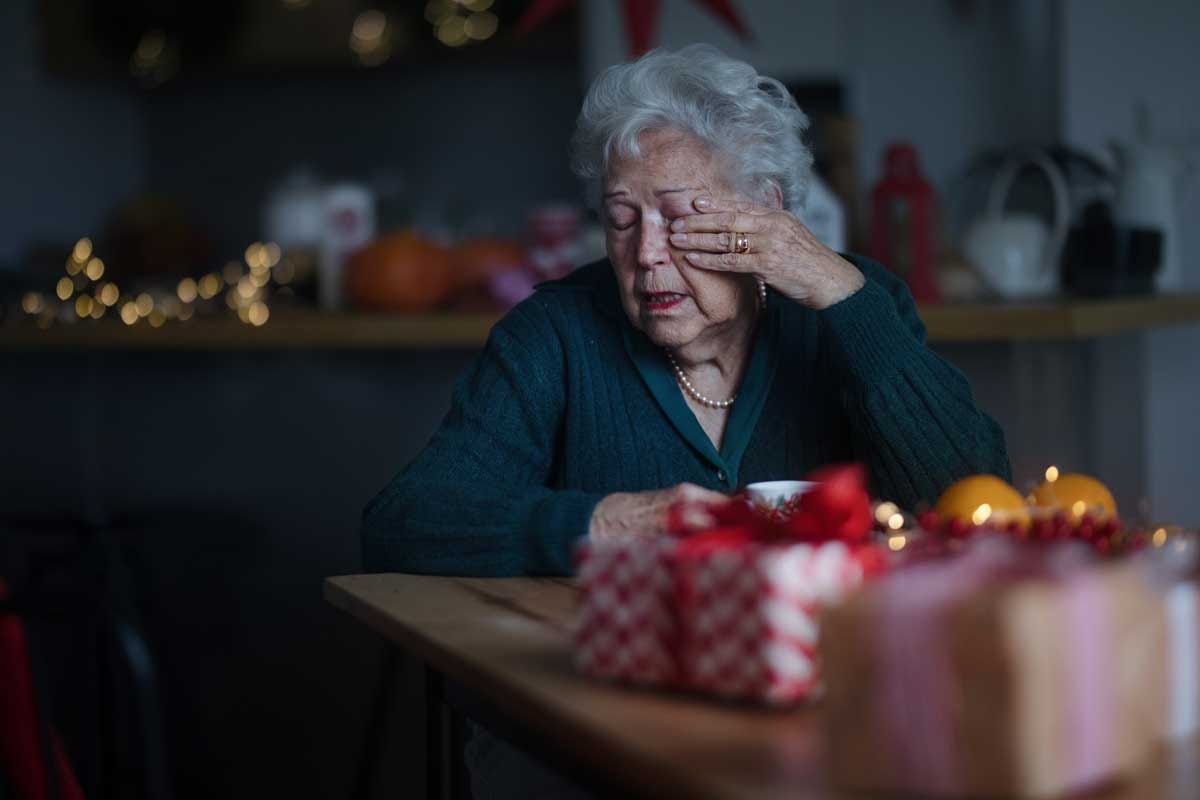 Elder woman looking despondent while sitting at kitchen table full of holiday gifts.