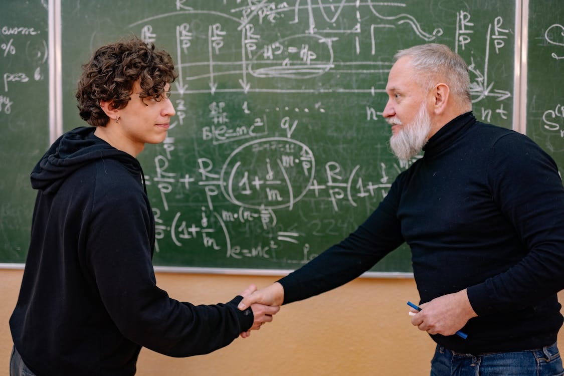 Free A professor and student shake hands in front of a chalkboard filled with mathematical formulas, symbolizing education and mentorship. Stock Photo